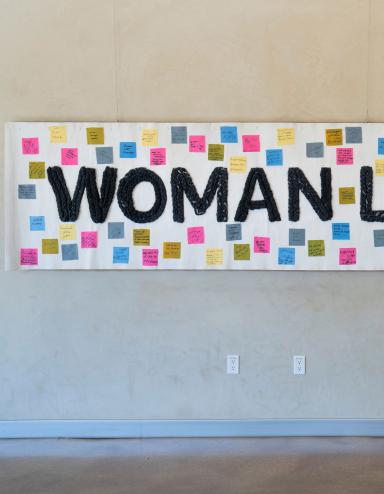 Une banderole blanche est accrochée à un mur, portant les mots "Femme, Vie, Liberté" en noir. De petits carrés de couleur avec du texte sont attachés à la bannière autour du texte. Visibilité masquée.