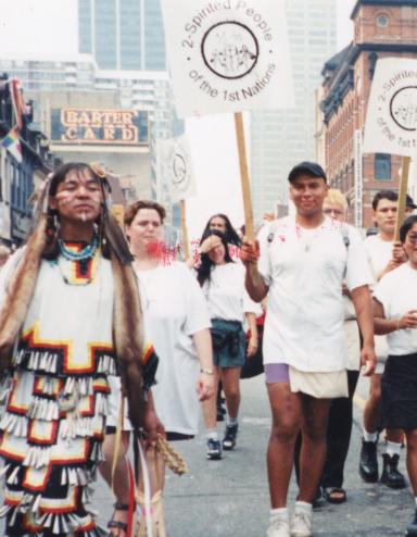 Un groupe de personnes portant des pancartes sur lesquelles on peut lire « 2-Spirited People of the 1st Nations » marchent au centre d’une rue de la ville, tandis que des gens regardent derrière des barrières de part et d’autre de la rue. Visibilité masquée.