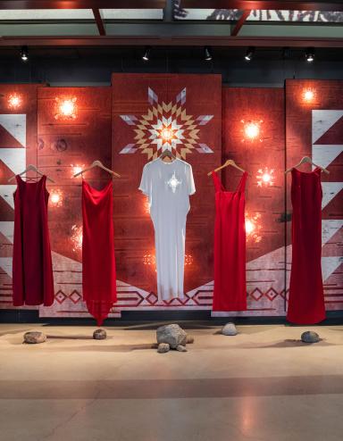 Six robes rouges et une robe blanche au milieu sont suspendues à des cintres en bois devant un arrière-plan. Une étoile blanche brille à l’emplacement du cœur sur la robe blanche centrale. L’arrière-plan présente des motifs sur une texture d’écorce de bouleau. Certains motifs sont éclairés par derrière pour créer des effets lumineux ressemblant à des étoiles sur les panneaux. Visibilité masquée.