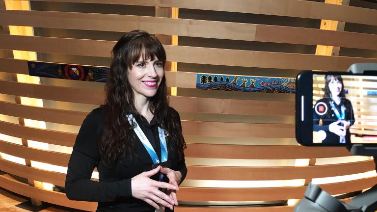 Une femme souriante à la peau claire se fait filmer avec un téléphone portable. Elle porte un cordon autour du cou sur lequel on peut lire "Laissez-vous inspirer" et se tient devant un élément d'exposition fait de bandes de bois horizontales, certaines décorées d'art autochtone. Visibilité masquée.