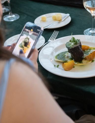 Une femme à la peau claire et aux longs cheveux bruns utilise son téléphone portable pour prendre une photo d’une assiette de nourriture gastronomique posée sur une table. Visibilité masquée.