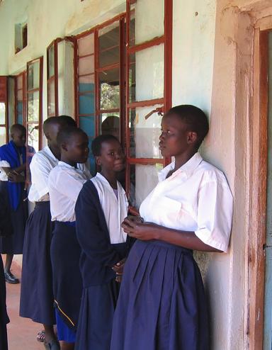 A group of young women and girls in their teens stand together on the front porch of a building. Most are dressed in white blouses and blue skirts, with two girls also wearing blue sweaters. To the right, a doorway reveals a roomful of students sitting at wooden desks and writing on notepads. Visibilité masquée.