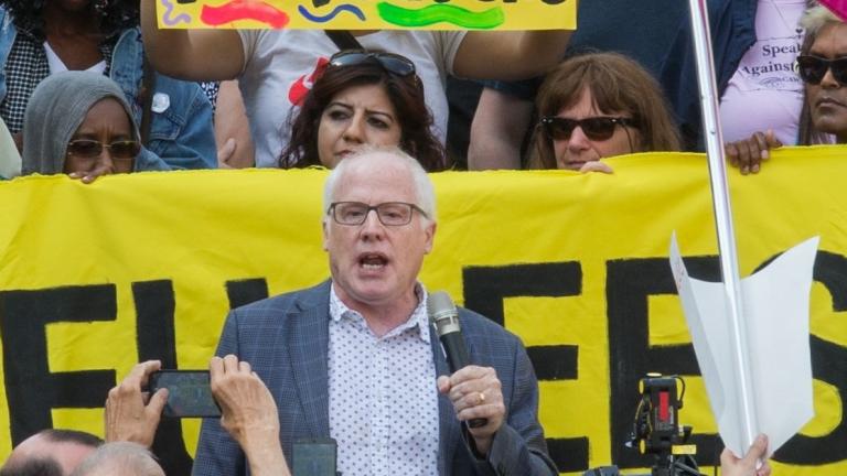 Un homme blanc aux cheveux blancs, portant des lunettes et un blazer, parle au micro. Il est entouré d'une foule de personnes brandissant des appareils photo, des pancartes de protestation et une banderole. Visibilité masquée.